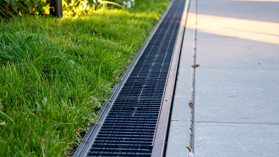 Black channel drain installed between a concrete driveway and green lawn for water drainage.