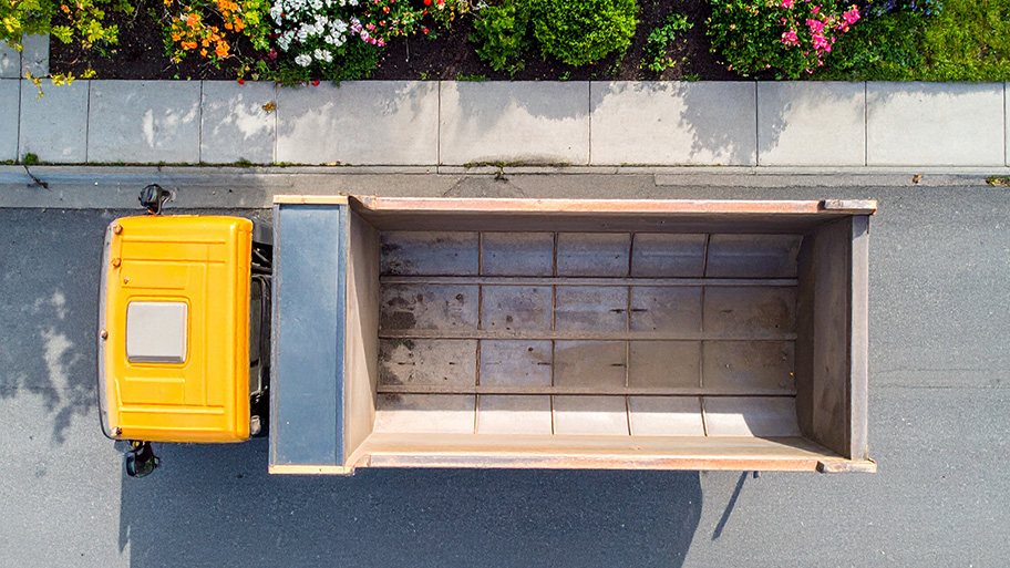 An aerial view of a bright yellow dump truck parked on an asphalt street with a sidewalk and planter box