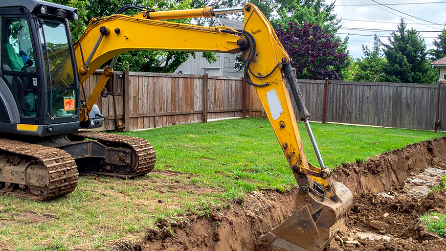 A yellow mini-excavator digs a trench in a residential backyard next to a wooden fence
