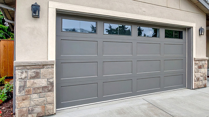 A light gray garage door with windows is set into a tan stucco house with stone accents and a concrete driveway