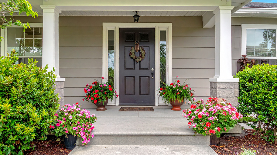A symmetrical front porch features a dark brown door with a wreath, flanked by glass panels, white columns, and vibrant red and pink flowers
