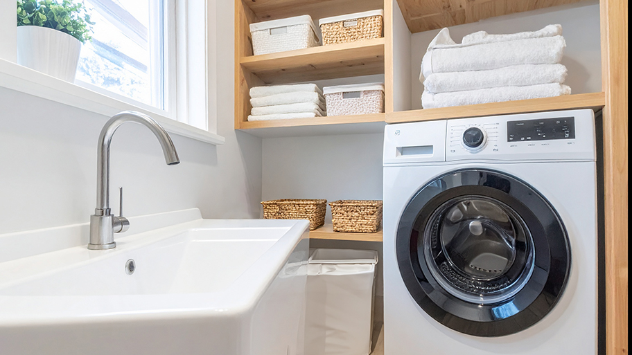 A modern laundry area in a garage with a white sink, a front-load washing machine, and wooden shelves holding towels and baskets