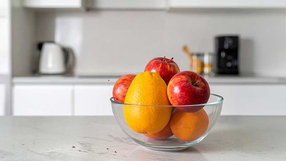 A glass bowl of apples and oranges sits on a kitchen counter, attracting small gnats or fruit flies