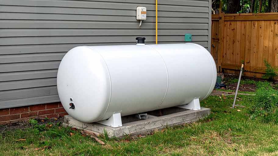 A large white propane tank rests on a concrete slab next to a gray house and a wooden fence.