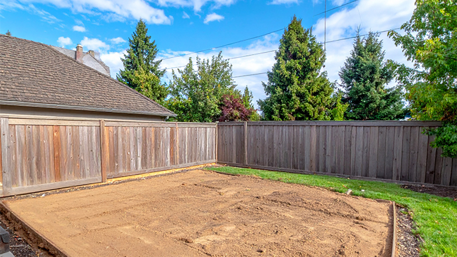 A backyard with a newly leveled area of packed dirt and a surrounding wooden fence and green grass.