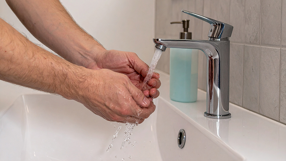 A person's hands are being washed under a stream of water from a modern chrome faucet in a white bathroom sink.