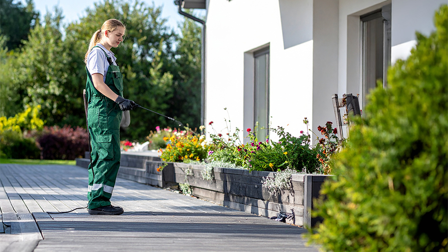 A professional in green coveralls sprays pesticide near a flower bed along the wooden deck of a modern home.
