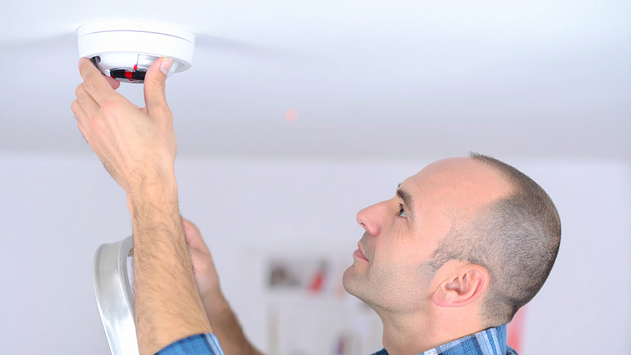 Man standing on a ladder replacing the battery in a ceiling-mounted smoke detector.