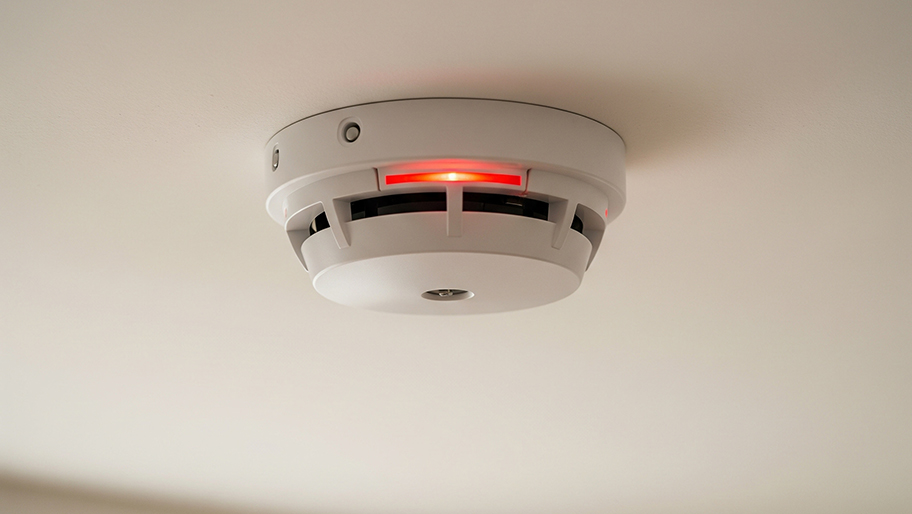 A ceiling-mounted smoke detector with a glowing red indicator light signals active status or alert mode.