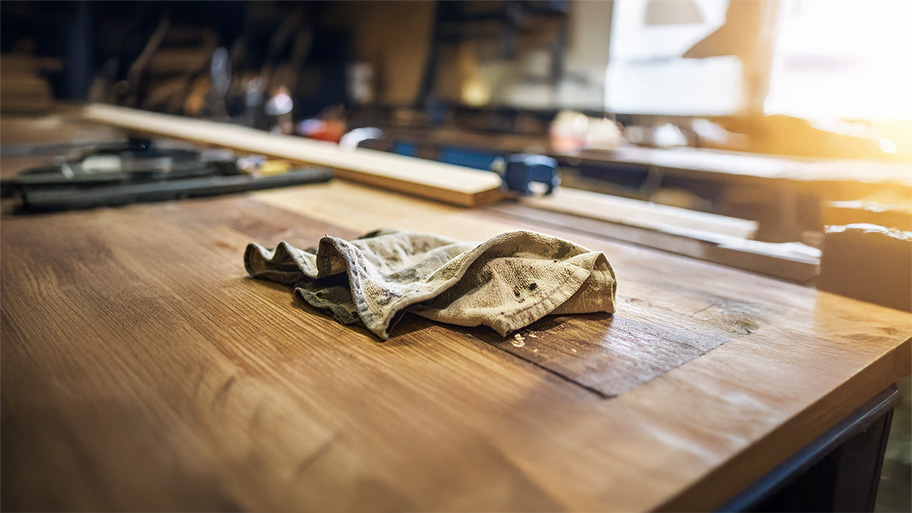 A hand in a blue glove wipes a glossy, dark wood floor with a yellow cloth.