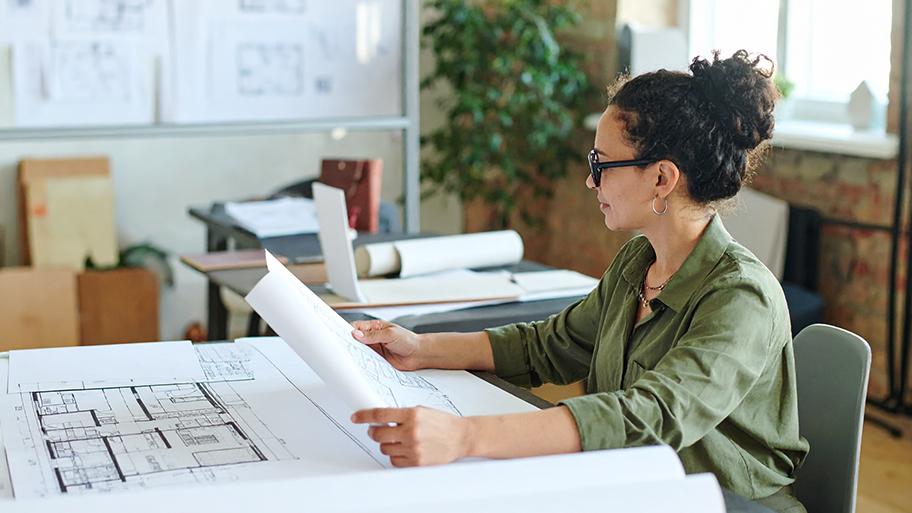 A professional female structural engineer with glasses examines blueprints at her desk in a modern office space.