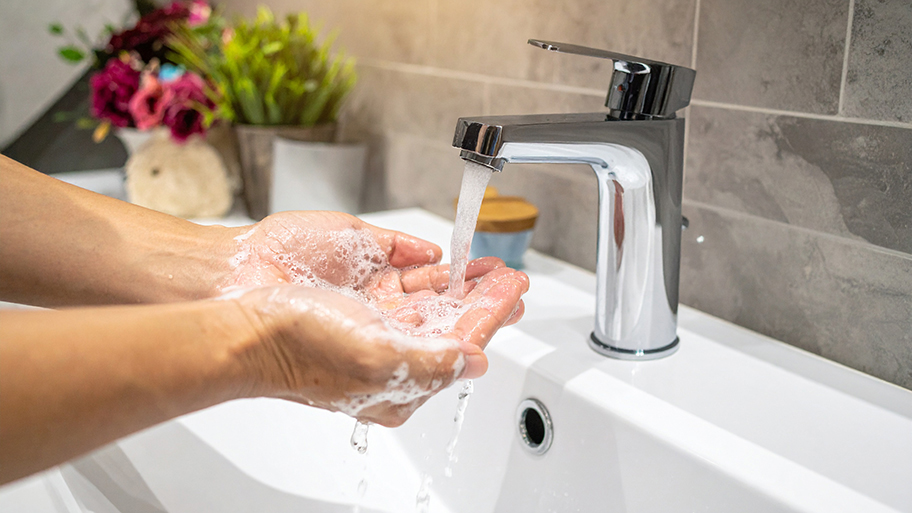 Soapy hands under a running faucet in a bathroom sink with decorative flowers in the background.