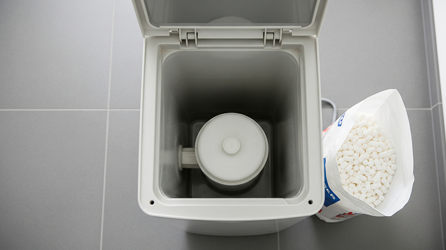 Opened water softener tank showing the resin chamber beside a bag of white salt pellets on a tiled floor.