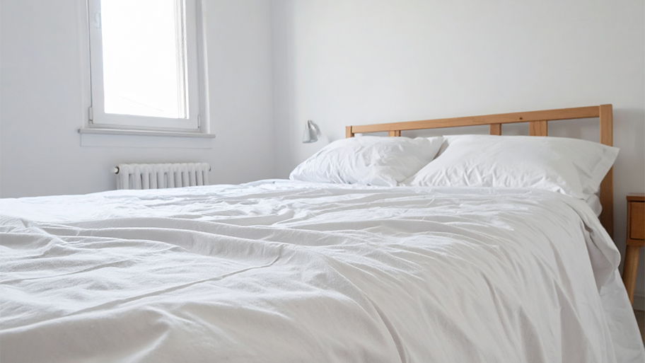 A simple wooden bed with a rumpled white duvet and pillows sits in a stark, all-white bedroom.