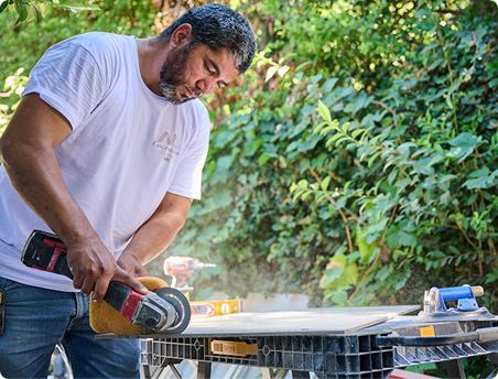 Home services pro using a power grinder to cut a tile outdoors, with dust flying as he works on a table surrounded by tools and greenery in the background.