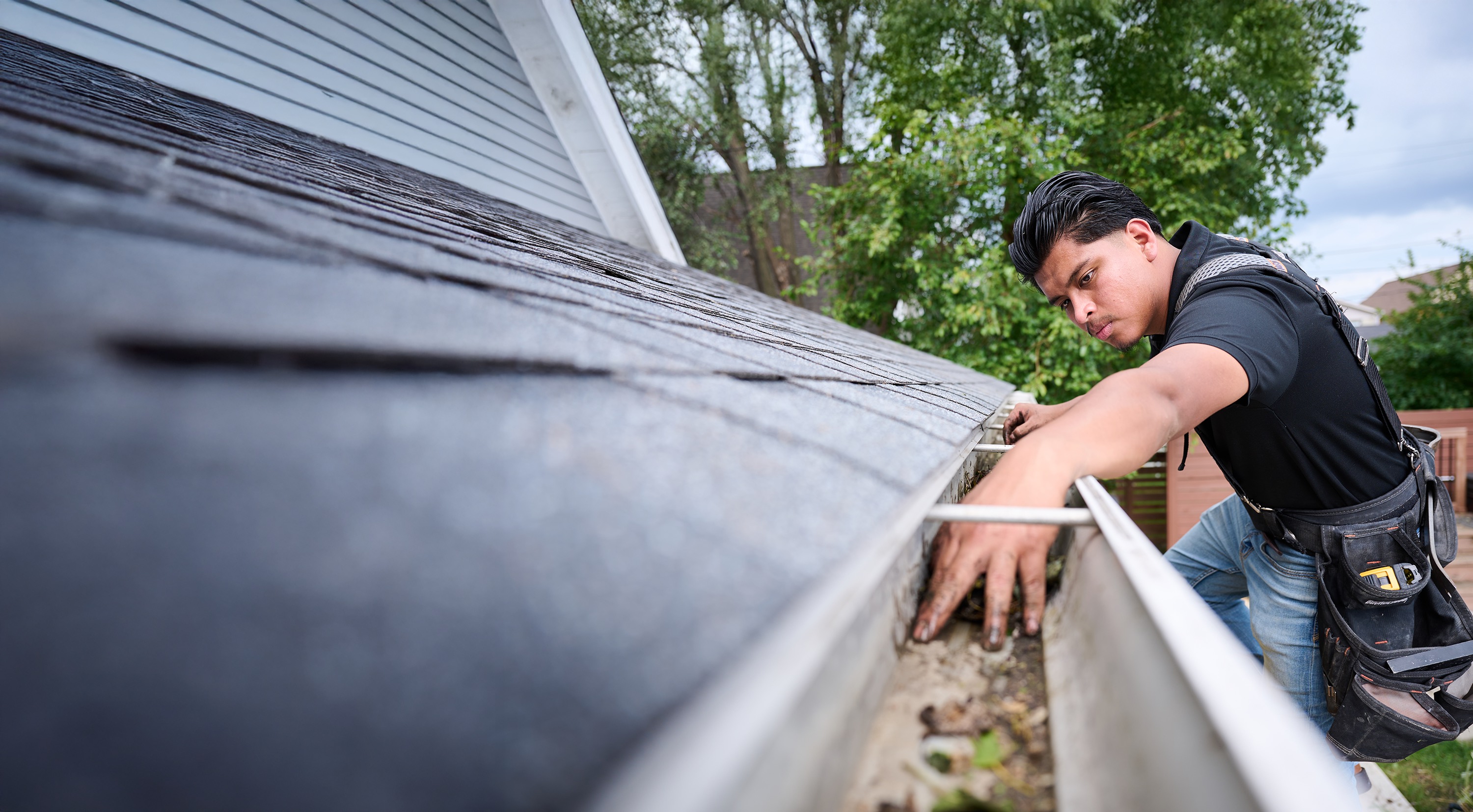 A professional cleans debris from a roof gutter while standing on a ladder beside a house.