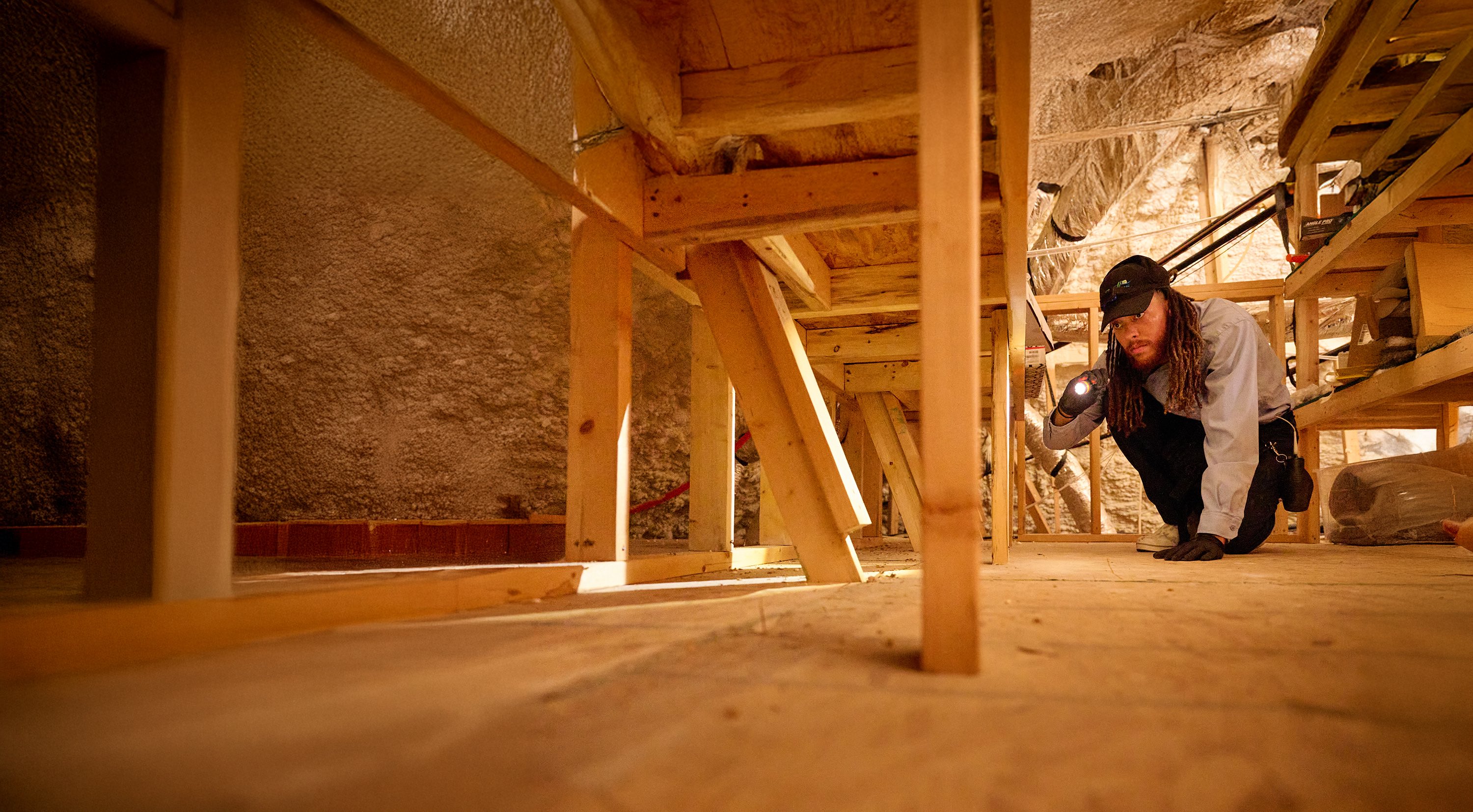 A pest control professional examines a crawl space under a home using a flashlight.