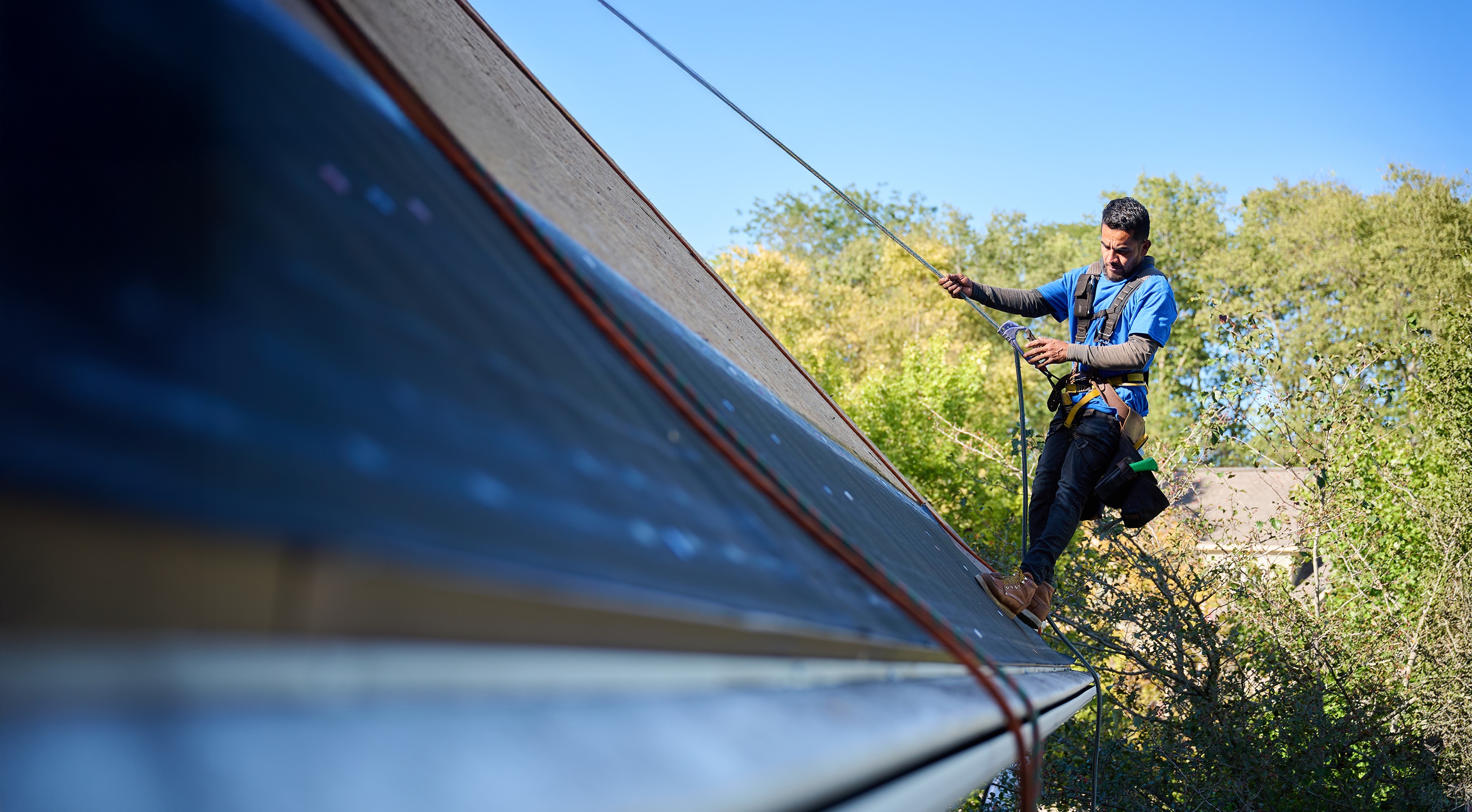 A roofer wearing safety gear works on a sloped roof using a harness and rope.