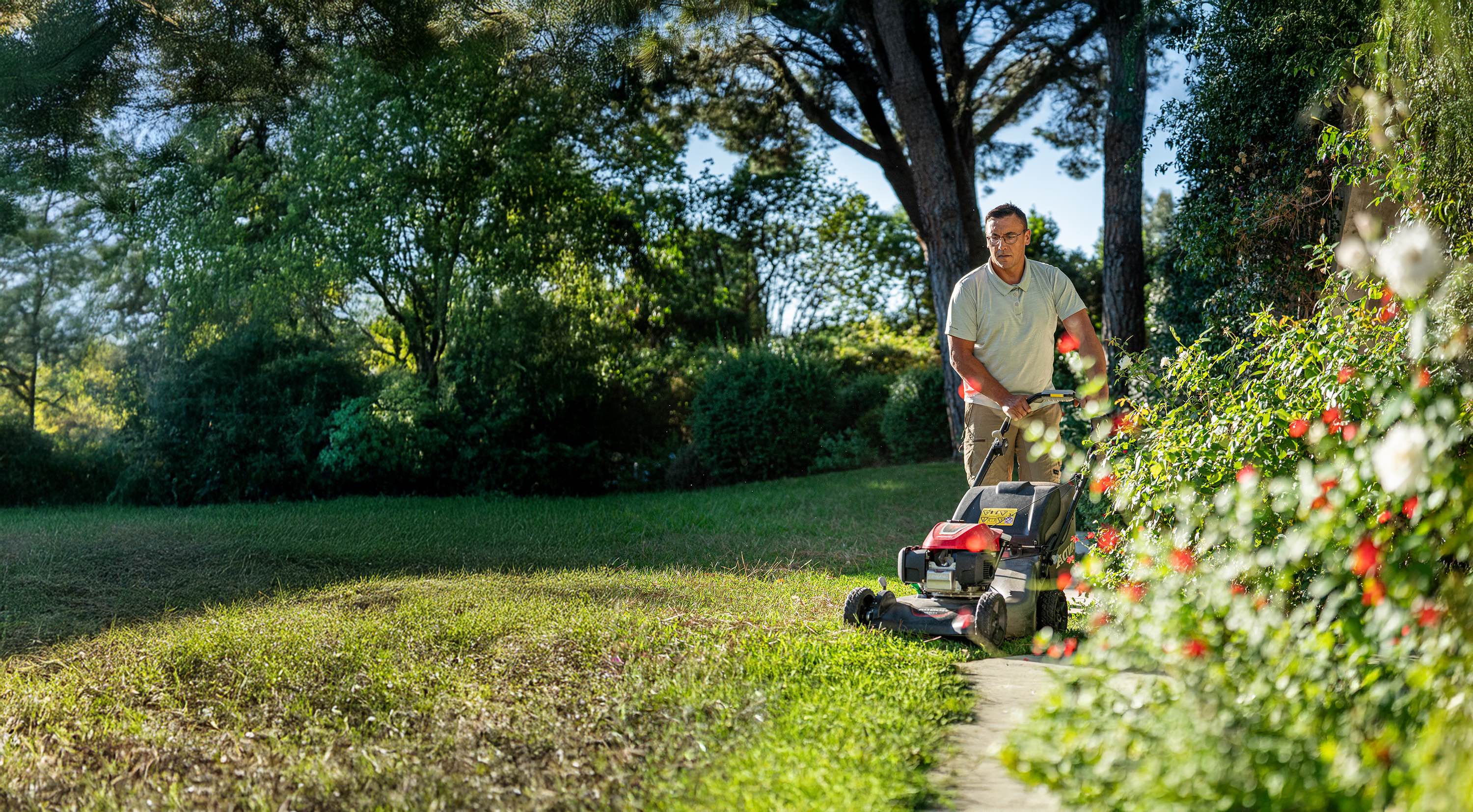 A landscaping professional pushes a lawn mower along a garden path, trimming grass beside shrubs and trees.
