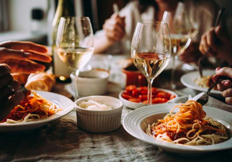 Middagsbjudning med pasta i tomatsås och vitt vin i glas på bord.