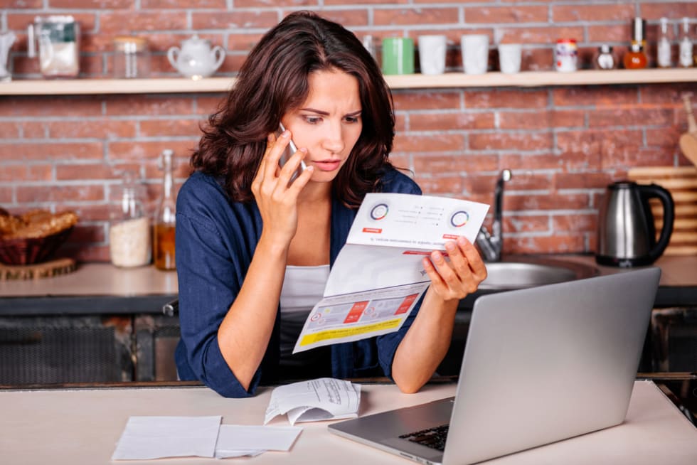 woman sitting in the kitchen with dissatisfied face in front of laptop holding utility bills, talking on mobile phone