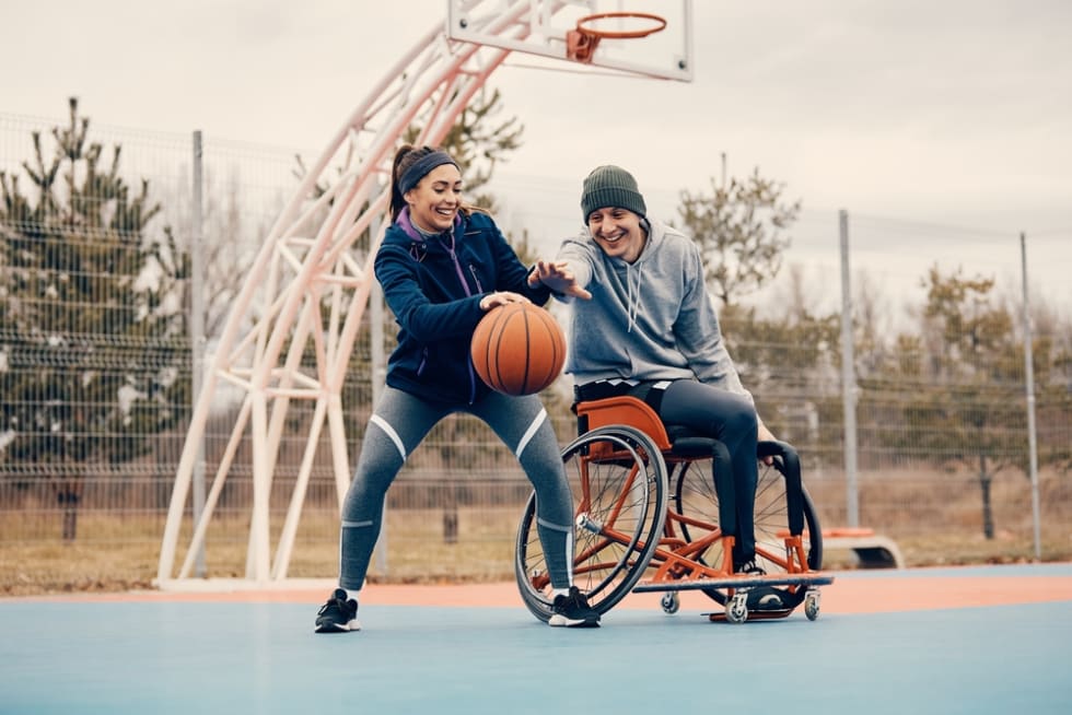 Happy athlete in wheelchair and his female friend having fun while playing basketball outdoors