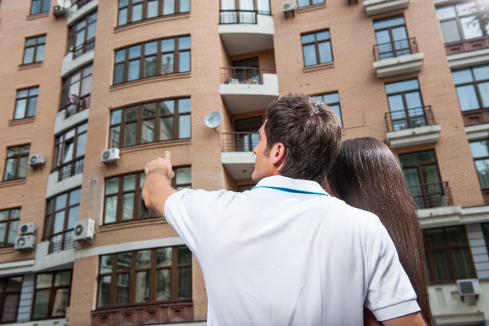 couple standing with their backs towards camera. man pointing to apartment window for brunette woman