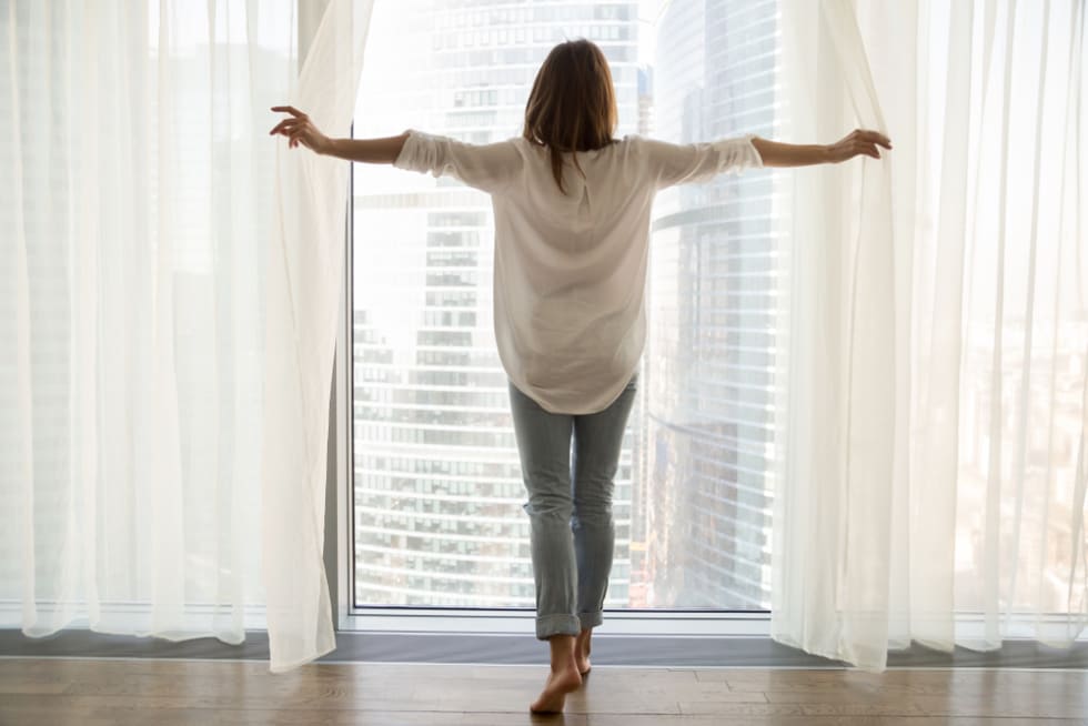 woman standing looking out of full-length window of luxury modern apartment or hotel room opening curtains in the morning enjoying sunlight and city skyscrapers
