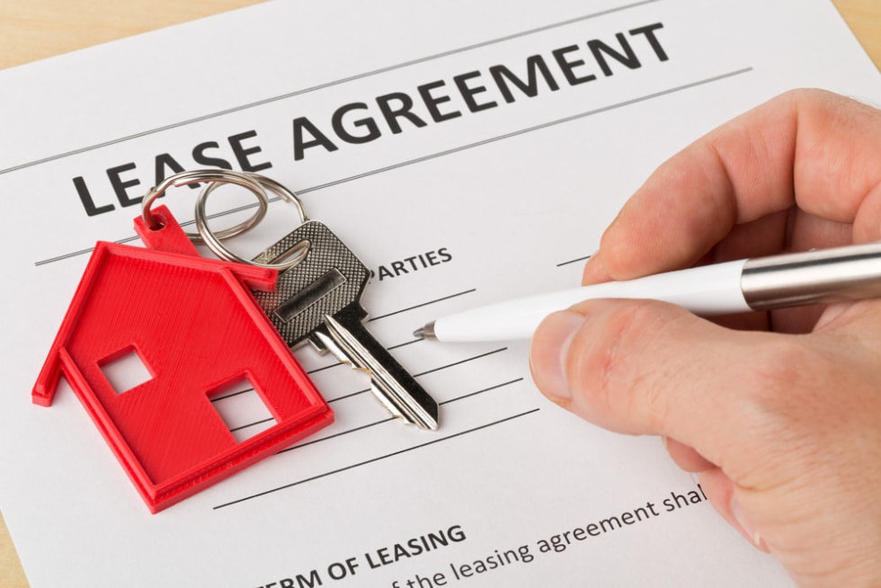  Man holding pen over house door key with red keychain pendant and lease agreement form on wooden desk - house or apartment rental concept