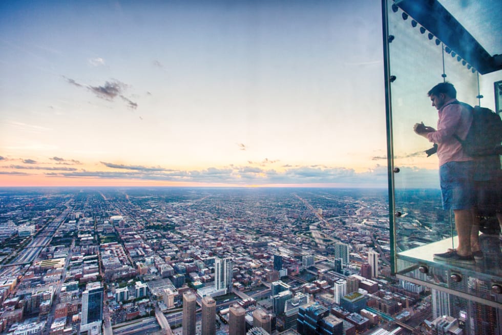 A tourist takes a picture of the Chicago skyline from one of the glass boxes of the Willis Tower Skydeck Ledge near sunset