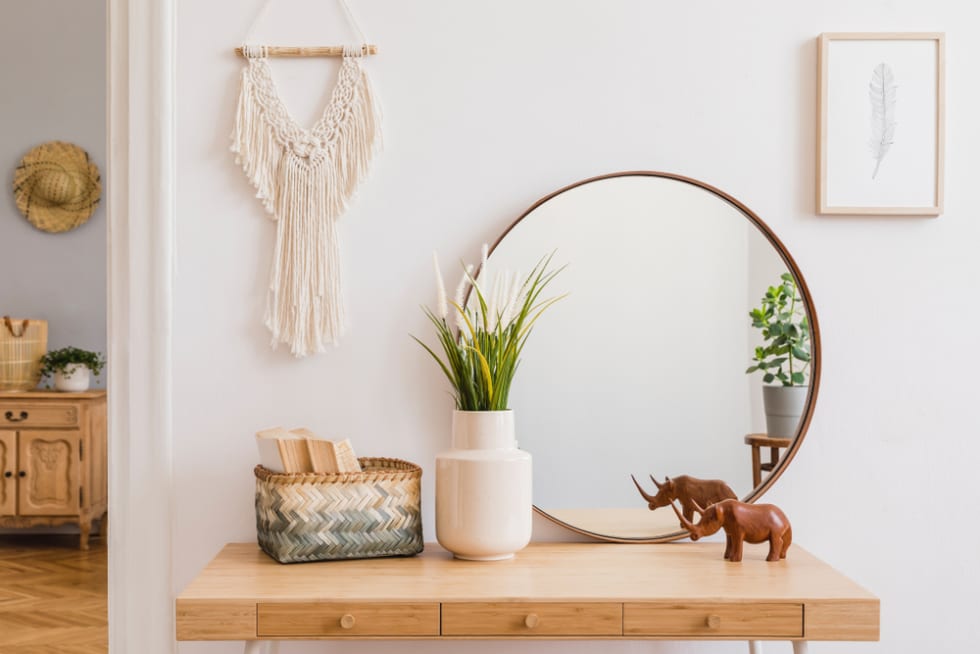 Sunny boho interiors of apartment with mirror, dressing table, furnitures, flowers, plants, rattan box, books, sculpture, macrame and design accessories.