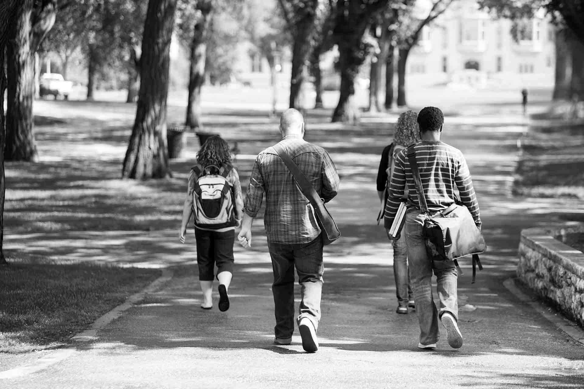 Four university students walking through a park holding textbooks and carrying backpacks.