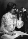 Portrait of Helen Keller sitting with a braille book and smelling flowers sitting in a vase beside her.