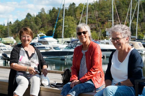 Tre damer som sitter sammen på en brygge med masse båter i bakgrunnen på sommerstid.