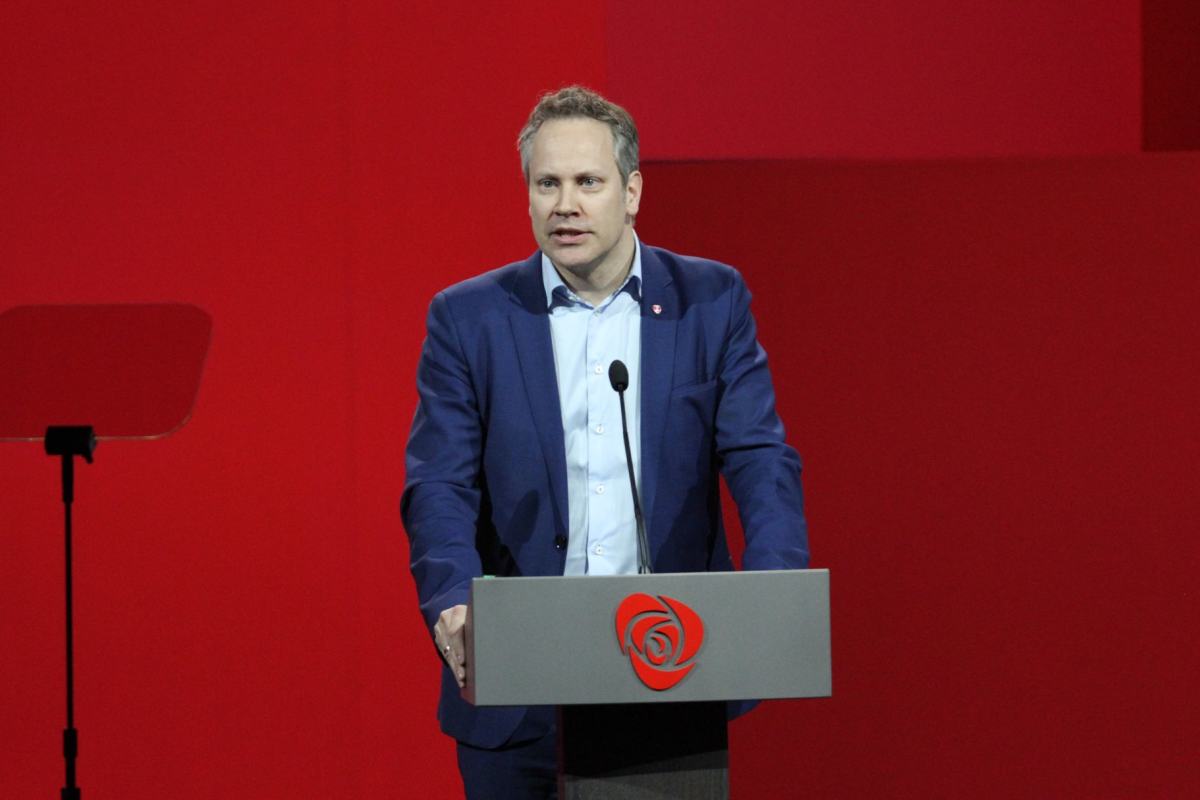 Mann på talerstol med blå dress og rød vegg bak