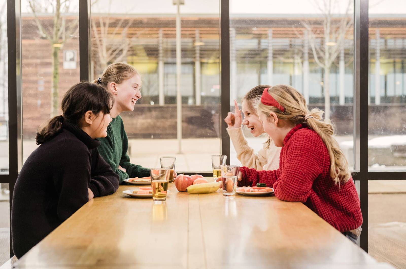 Fire smilende unge jenter sitter i et lyst lokale rundt et bord med mat på asjetter, frukt og glass med drikke.