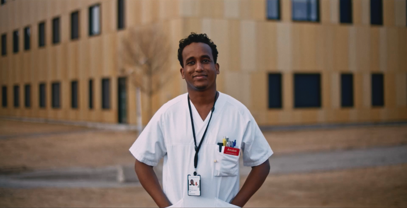 Mann i hvit sykepleieruniform, mørkt hår. Står utendørs.