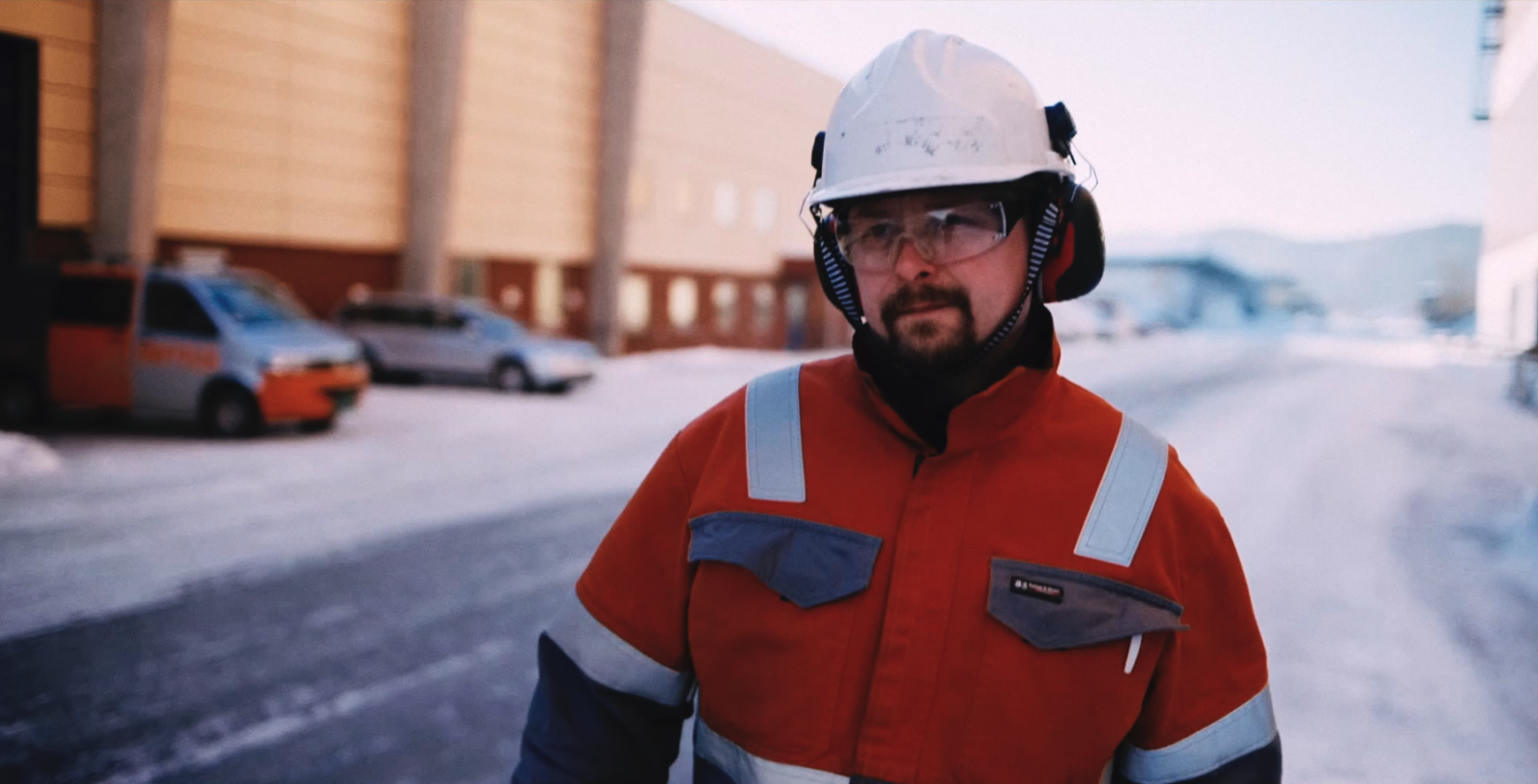 Mann i hvit hjelm og orange kjeledress, går bortover. Utendørs.
