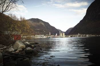 Oversiktsbilde av en bygd i enden av en fjord, industribygg ruver høyt foran fjellene, i forgrunnen ligger havet stille. Foto: Øivind Haug