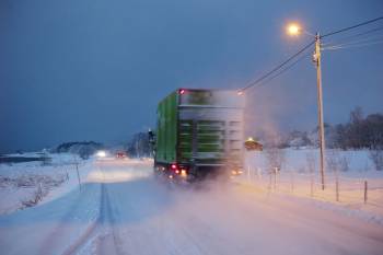 En grønn lastebil kjører langs en snøkledd vei, snøen spruter opp bak den. Foto: Øivind Haug