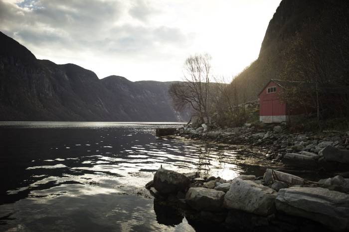 Et rødt naust ligger ved kanten av en fjord, omgitt av fjellvegger. Foto: Øivind Haug