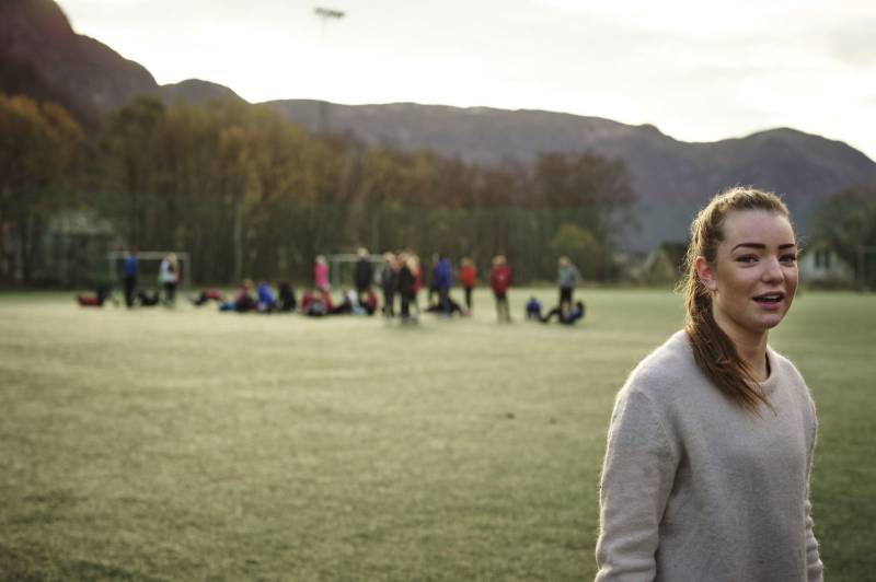 I forgrunnen ser vi en jente, i bakgrunnen står et fotballlag på banen. Foto: Øivind Haug