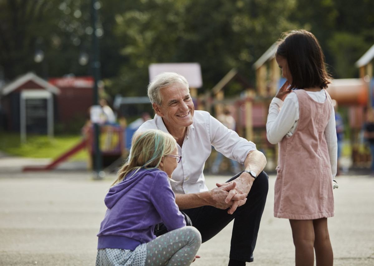 Jonas Gahr Støre sitter på huk i en skolegård og snakker med to jenter på skolefritidsordningen (SFO).