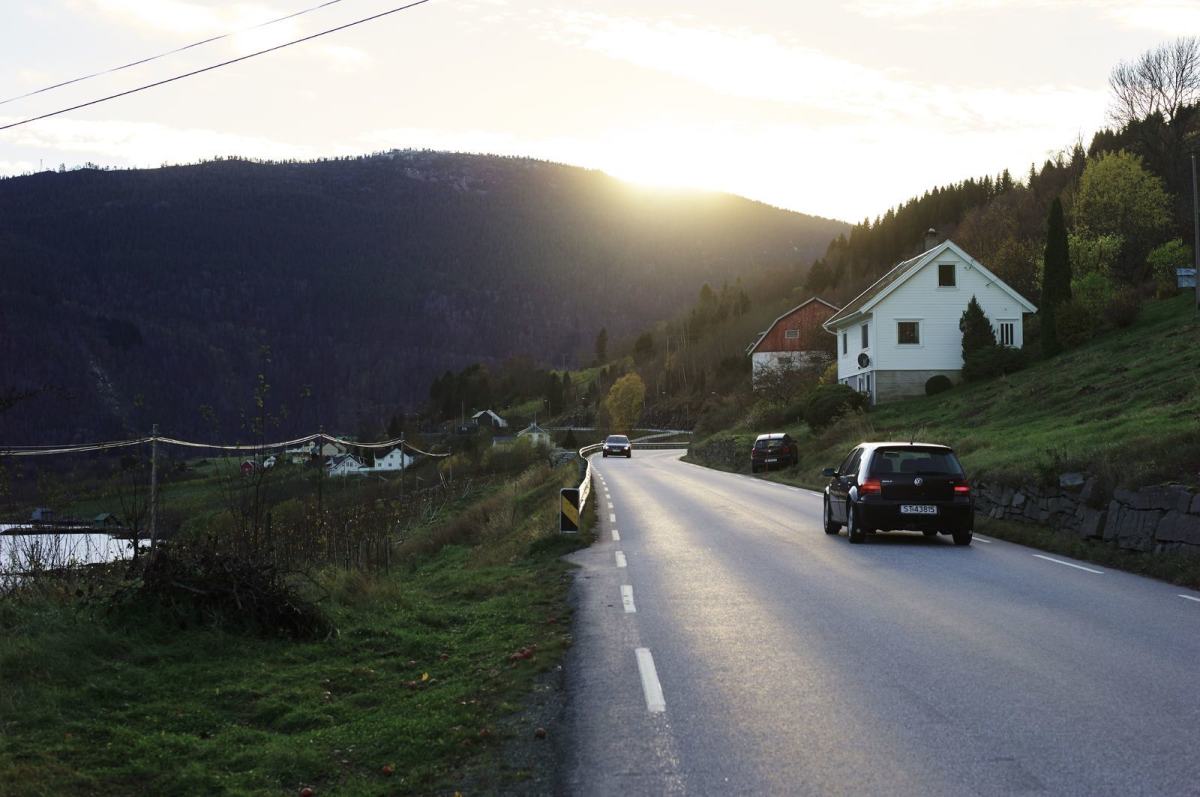 En landevei strekker seg gjennom landskapet, i bakgrunnen går sola ned bak et fjell. Foto: Øivind Haug