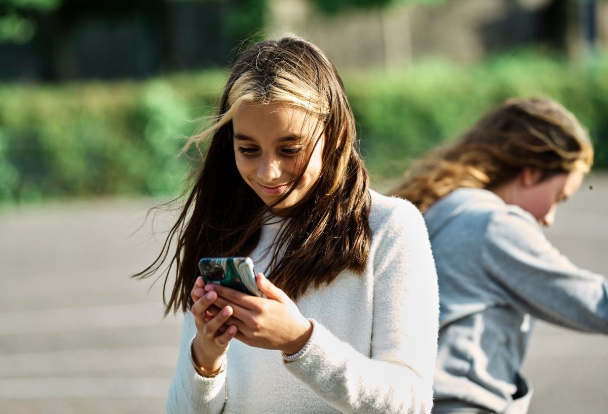 En ung jente står og ser på mobilen sin mens en annen jente leker i unnen.