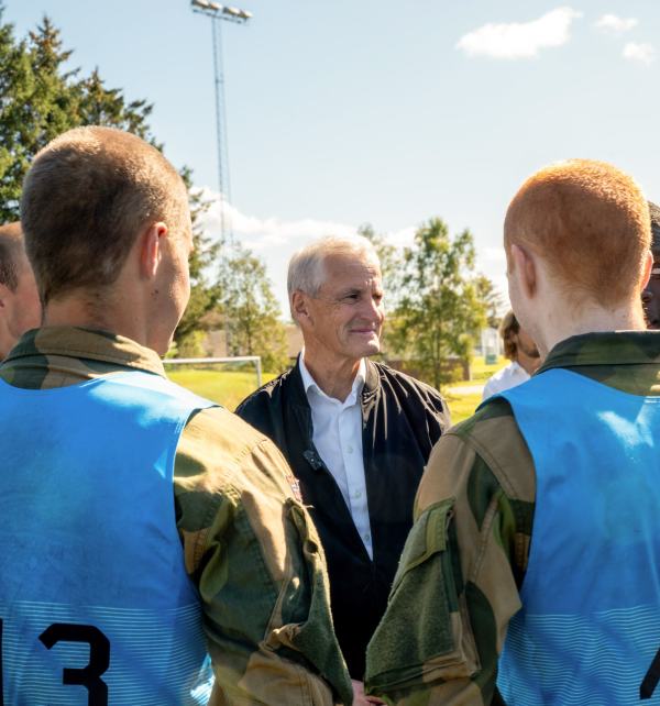 Statsminister Jonas Gahr Støre står og snakker med flere soldater ikledd forsvarsuniform og blå vester utendørs.