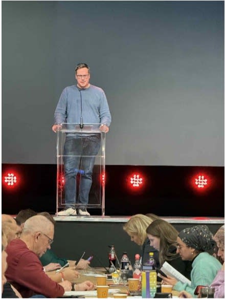 Karl Henrik Storhaug Reinås taler til representantskapet i Oslo AP fra en scene med gode partikamerater på et bord foran i salen.