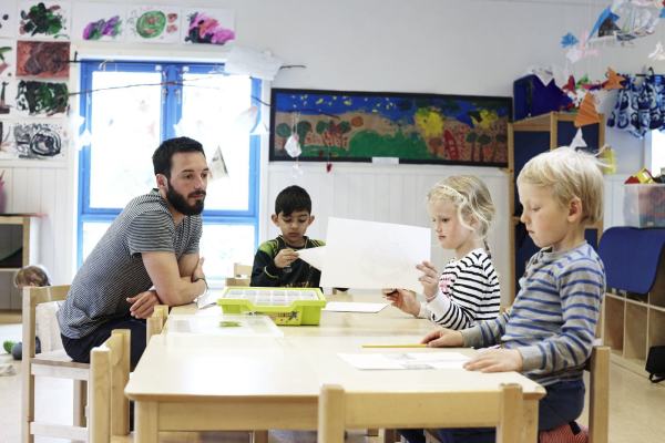 Tre barn og en ansatt sitter rundt bordet, det ene barnet viser fram en tegning hun har laget. Foto: Øivind Haug