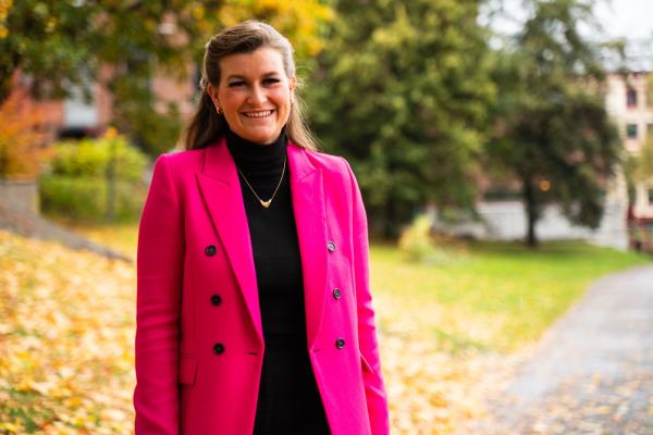 Thilde står i en rosa jakke foran noen grønne trær, og grønt gress. Det er noen bygg du så vidt ser i bakgrunnen