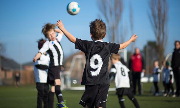 To gutter i forgrunnen, en i svart fotballskjorte og den andre i hvit og svar header fotball til hverandre. Flere gutter som spiller fotball i bakgrunnen.